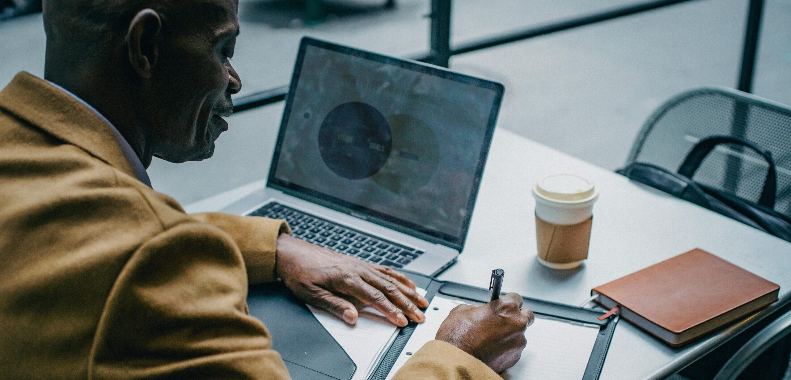 Person working outdoors with a laptop showing a goals-and-action diagram, writing notes on a clipboard beside a coffee cup.
